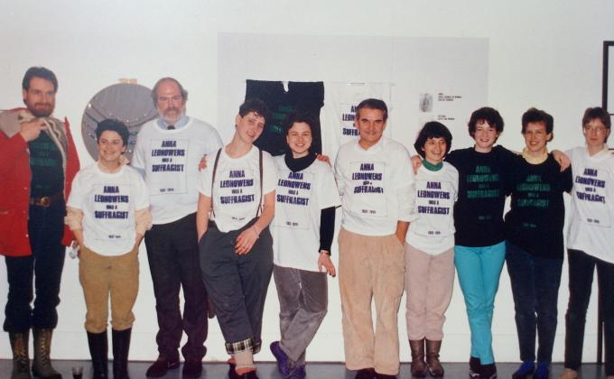 Ten people of varying heights and genders stand in front of a gallery installation. Each one is wearing an oversized white or black t-shirt on top of their other clothes which reads ANNA LEONOWENS WAS A SUFFRAGIST in purple or green capital lettering. Cathy Busby stands in the middle of the line of folks smiling at the exhibition she and her staff put together.