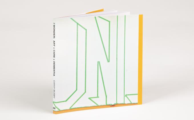 A small square book rests upright at an angle, the spine reads: I WONDER: ART + CARE + DEMENTIA CATHY BUSBY AM. The cover of the book is white with abstract letter-looking outlines in green painters tape. 