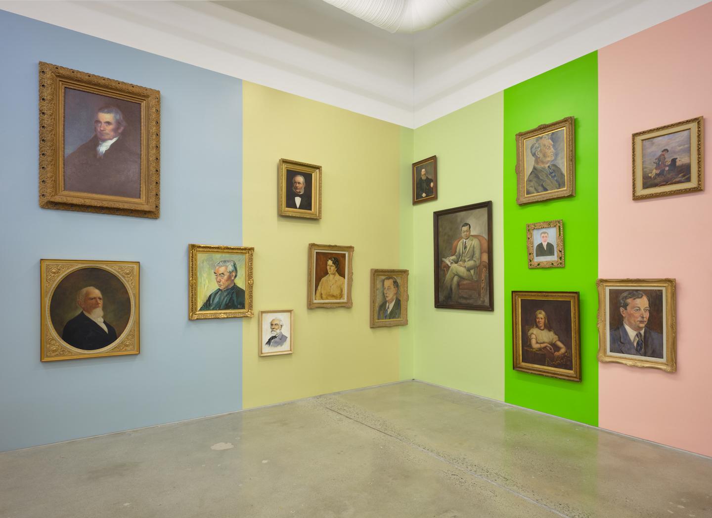 A gallery wall is painted in vertical blocks of dusty blue, wheaty yellow,  pale green, acid green and light pink providing the backdrop for a series of portraits with antique style wooden frames. The portraits rest at various heights and proximities; some are hung against just one colour while others sit on the border where two blocks meet. Each block of colour is reflected in the adjacent portrait’s colouration. 