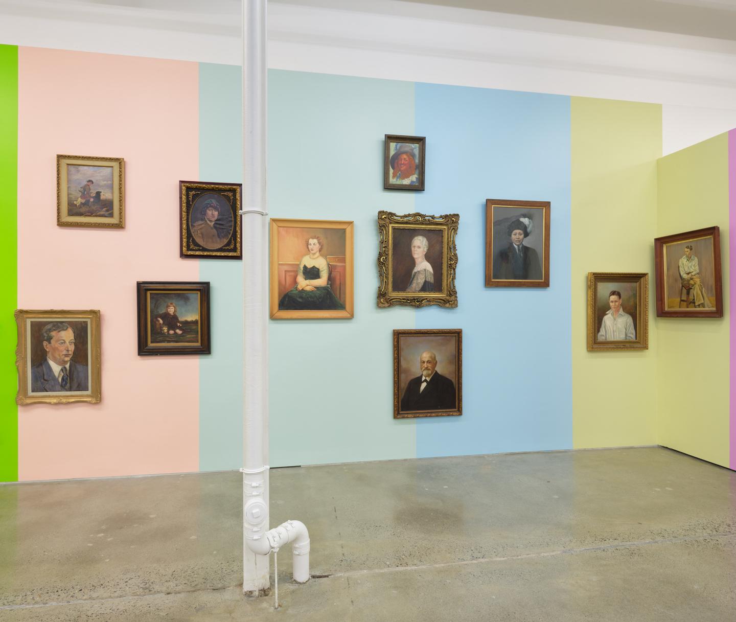 A gallery wall is painted in vertical blocks of pale pink, baby blue, eggshell blue, and pale yellow providing the backdrop for a series of portraits with antique style wooden frames. The portraits rest at various heights and proximities; some are hung against just one colour while others sit on the border where two blocks meet. Each block of colour is reflected in the adjacent portrait’s colouration. 