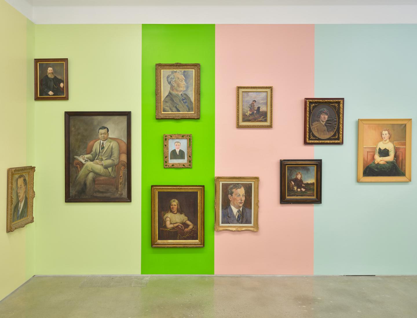 A gallery wall is painted in vertical blocks of pale yellow, pale green, acid green, pale pink and baby blue providing the backdrop for a series of portraits with antique style wooden frames. The portraits rest at various heights and proximities; some are hung against just one colour while others sit on the border where two blocks meet. Each block of colour is reflected in the adjacent portrait’s colouration. 