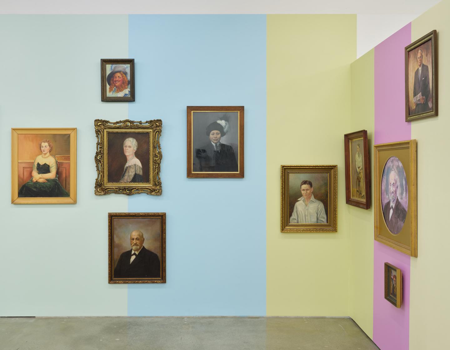 A gallery wall is painted in vertical blocks of baby blue, eggshell blue, pale yellow, light fuchsia and dusty yellow providing the backdrop for a series of portraits with antique style wooden frames. The portraits rest at various heights and proximities; some are hung against just one colour while others sit on the border where two blocks meet. Each block of colour is reflected in the adjacent portrait’s colouration. 