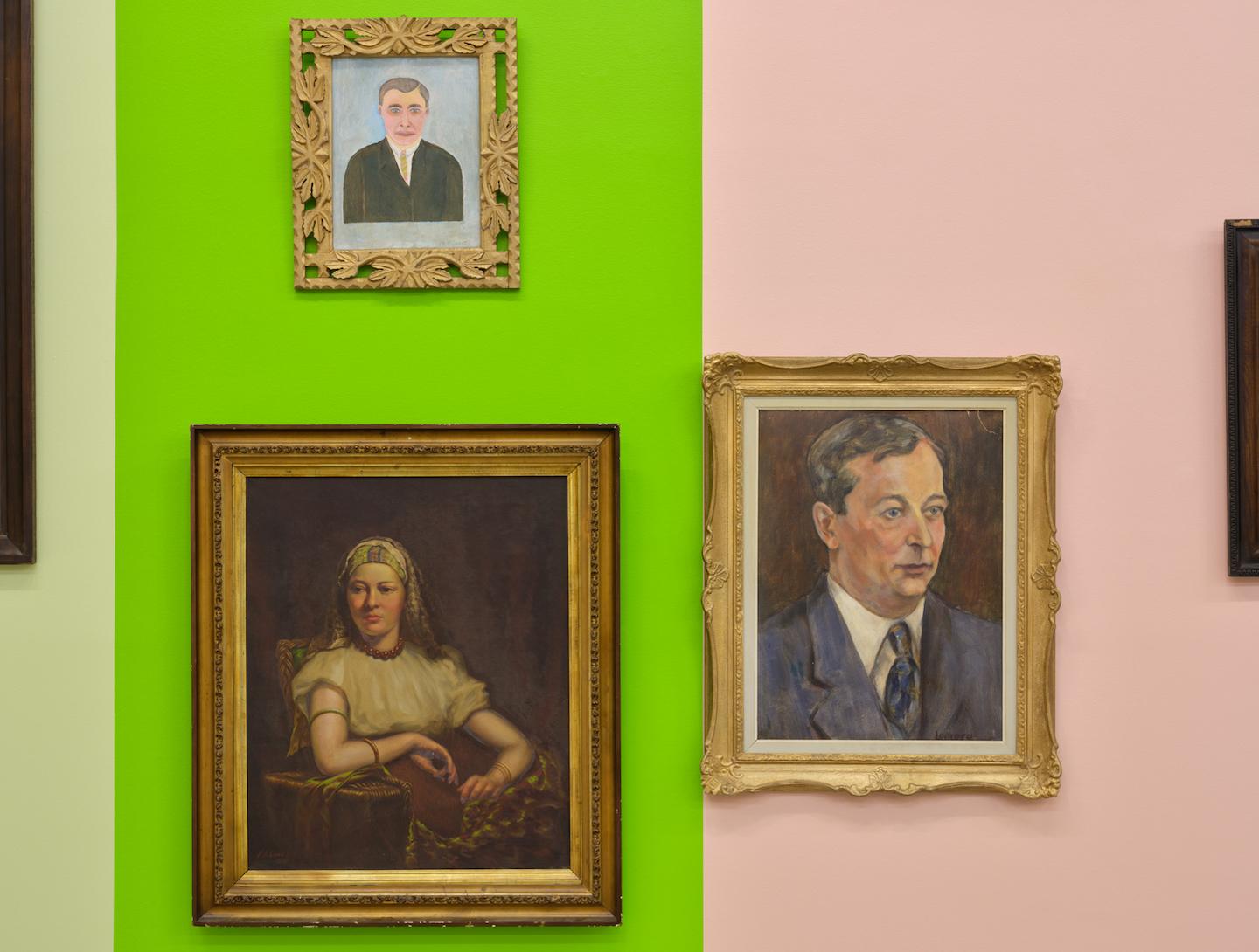 Three portraits are hung between acid green and pale pink wall panels. The green eyes of a pencil sketched man and the rosy cheeks of two pale skinned figures tone with the painted blocks of colour.