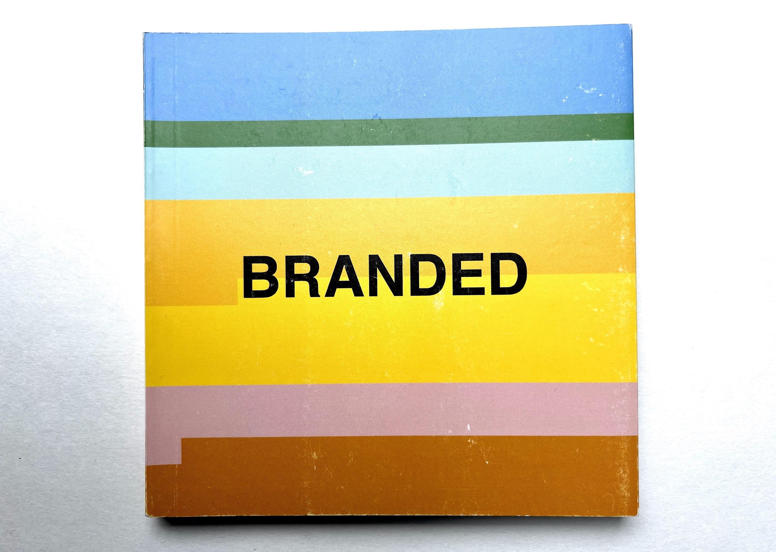 A small square book with multicoloured horizontal stripes in blues, greens and yellows rests on a white background. The word Branded is written in all caps in a black sans serif font in the center of the front cover.