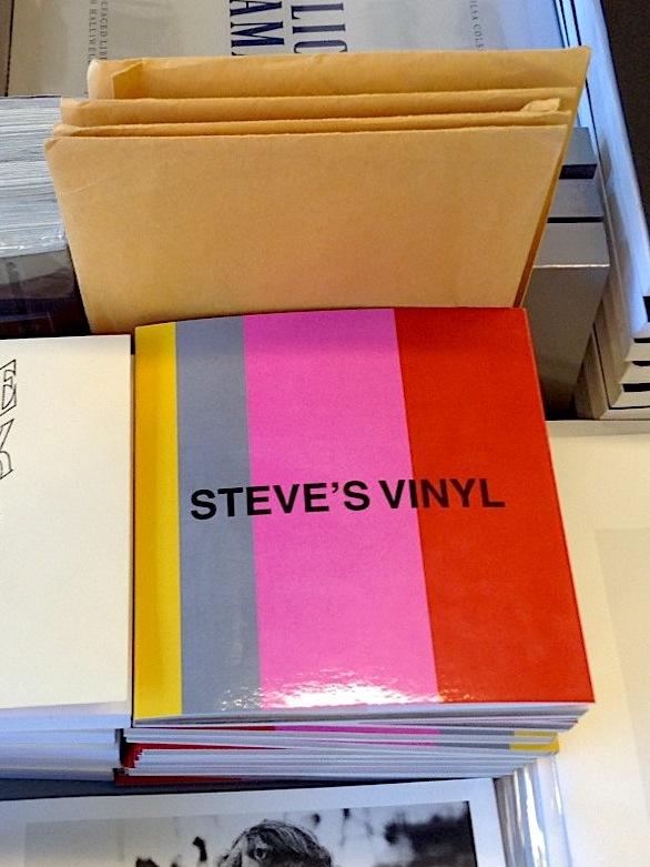 A stack of square books boldly striped with yellow, grey, pink and red with the title Steve’s Vinyl written in all caps in black font rests among other stacked publications on a table. Its glossy cover glints in the stark overhead lighting. The image is cropped tightly around this book so the surrounding publications are not discernible. 