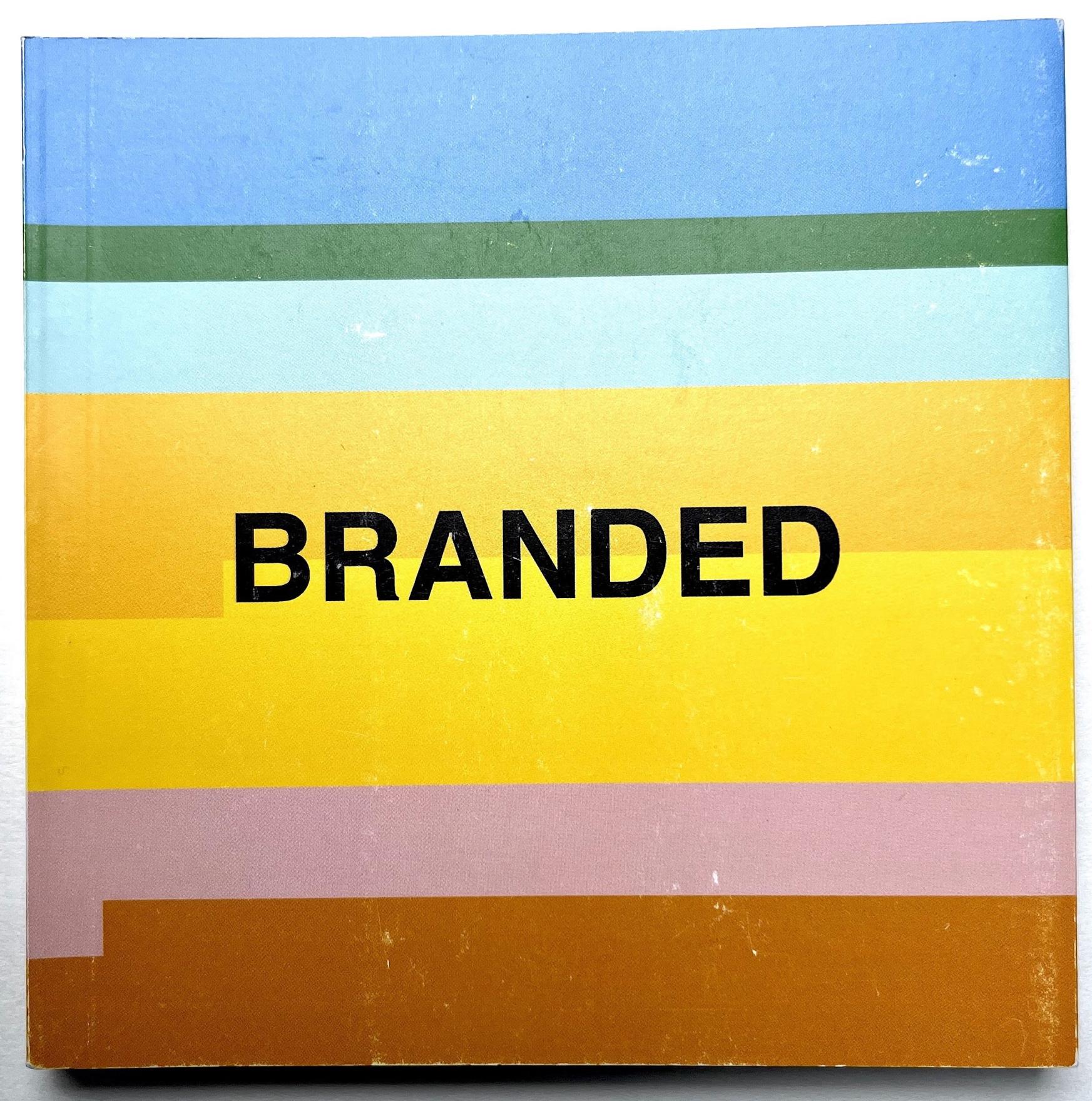 A small square book with multicoloured horizontal stripes in blues, greens and yellows rests on a white background. The word Branded is written in all caps in a black sans serif font in the centre of the front cover.