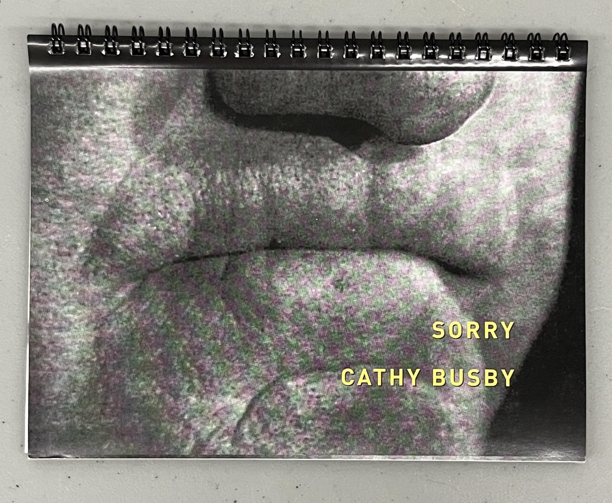 A black and white close up photo of a mouth with tightly pursed lips, perhaps holding back breath or biting back words, takes up the cover of the book. Sorry, Cathy Busby, is written in thick yellow letters near the right bottom corner. The book is bound by black metal ring binding and rests on a grey surface. 