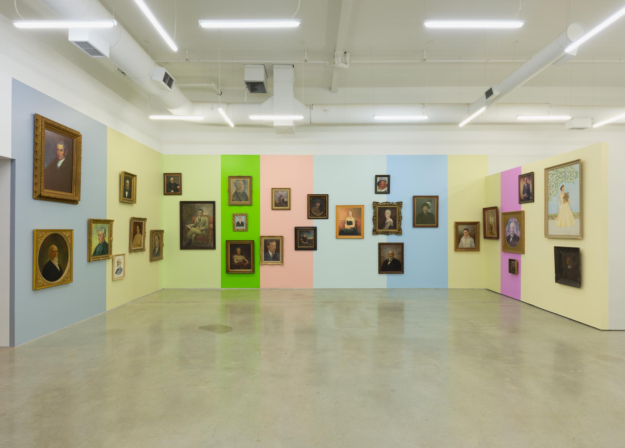 A gallery wall is painted in vertical blocks of pale pink, baby blue, eggshell blue, and pale yellow providing the backdrop for a series of portraits with antique style wooden frames. The portraits rest at various heights and proximities; some are hung against just one colour while others sit on the border where two blocks meet. Each block of colour is reflected in the adjacent portrait’s colouration. 