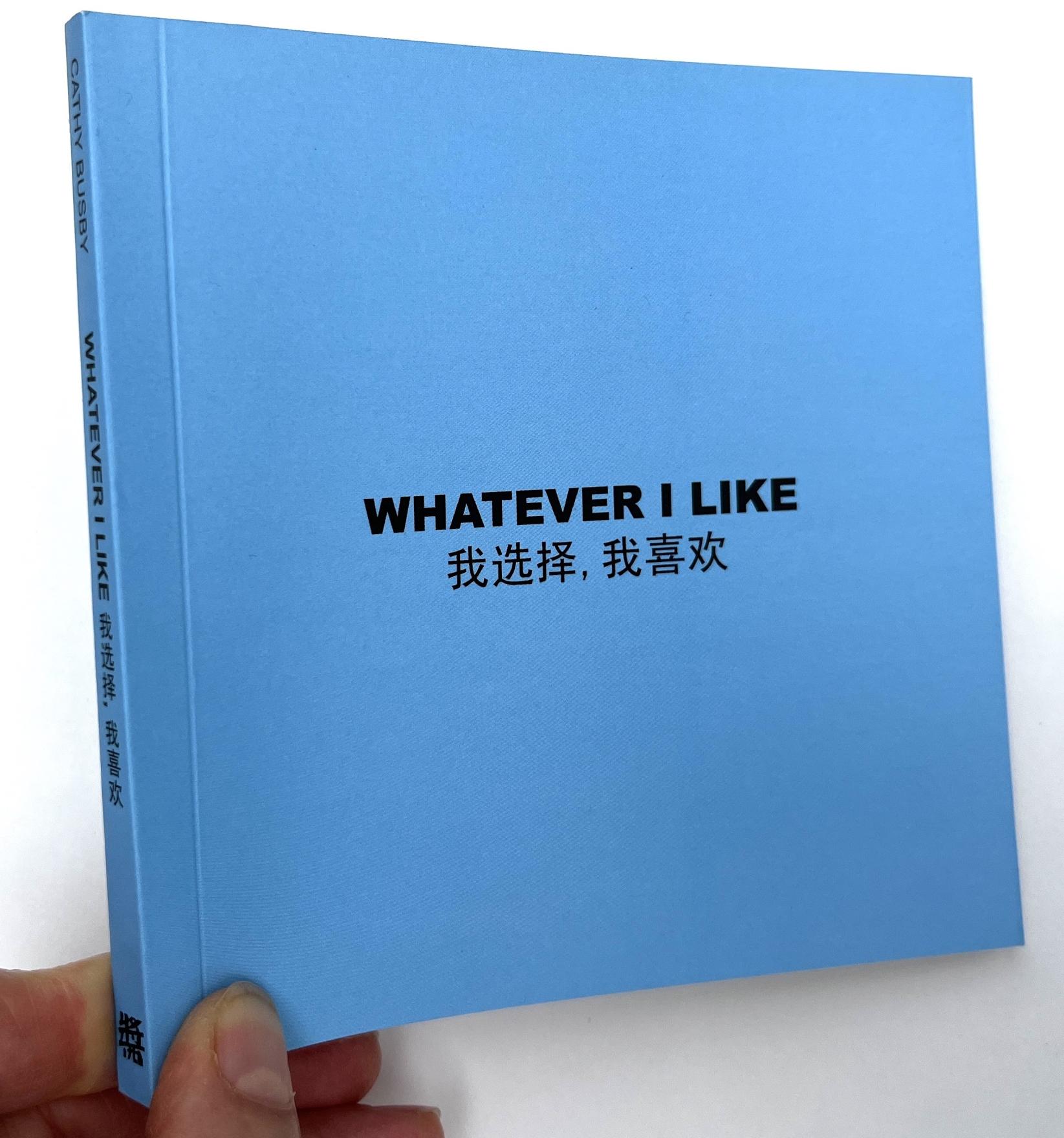 Fingers pinch and lift the bound corner of a small sky blue square paper bound book. Whatever I Like is written in the centre in black font in both English and Mandarin Chinese. At the angle the book is being tilted at you see this title is also written down the book’s blue spine after the artist's name: Cathy Busby. 