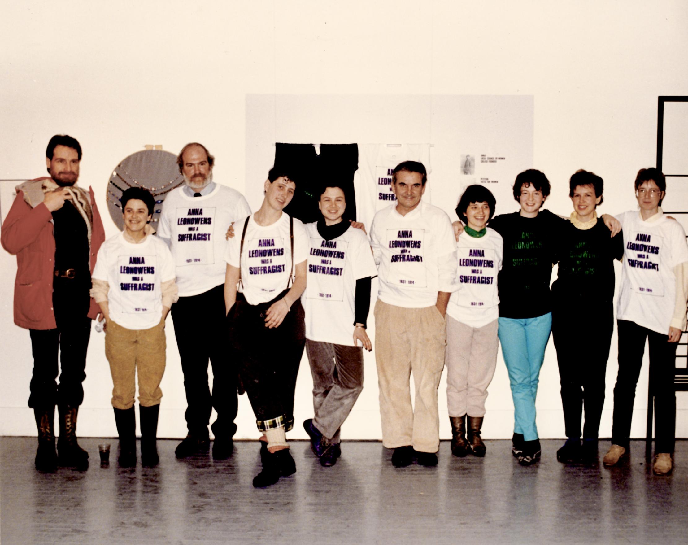 Ten people of varying heights and genders stand in front of a gallery installation. Each one is wearing an oversized white or black t-shirt on top of their other clothes which reads ANNA LEONOWENS WAS A SUFFRAGIST in purple or green capital lettering. Cathy Busby stands in the middle of the line of folks smiling at the exhibition she and her staff put together.