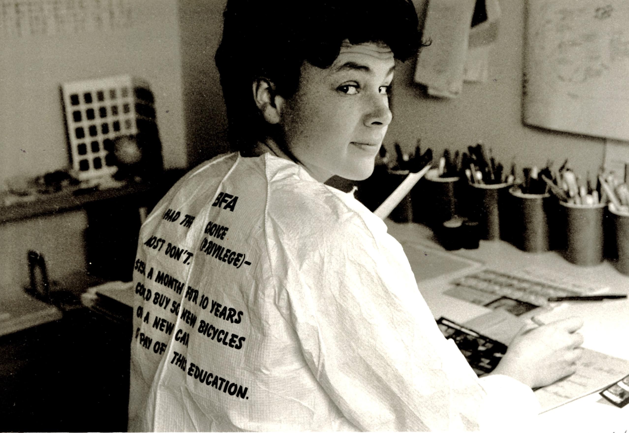 Cathy, with dark curly short hair wearing a paper painters’ suit, sits at a desk and looks over her shoulder at the camera. There is text printed on the back of the suit which reads: BFA, I had the choice (privilege) - most don't, $150 a month for 10 years could buy 50 new bicycles, or a new car, or pay off this education. The image is in black and white. 