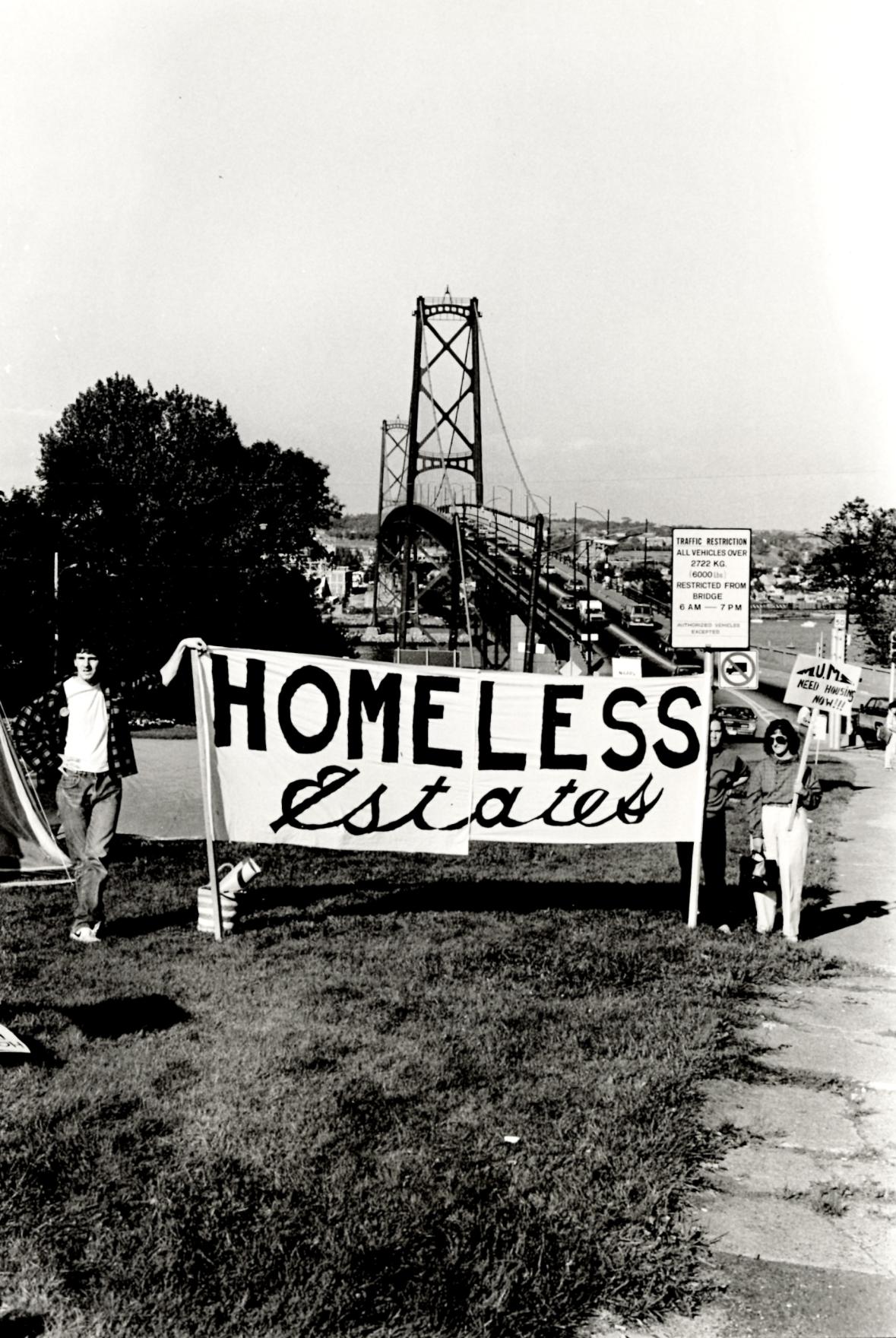 A black and white photograph shows three people standing by the roadside in a high traffic area holding up a large handmade banner which reads HOMELESS ESTATES.