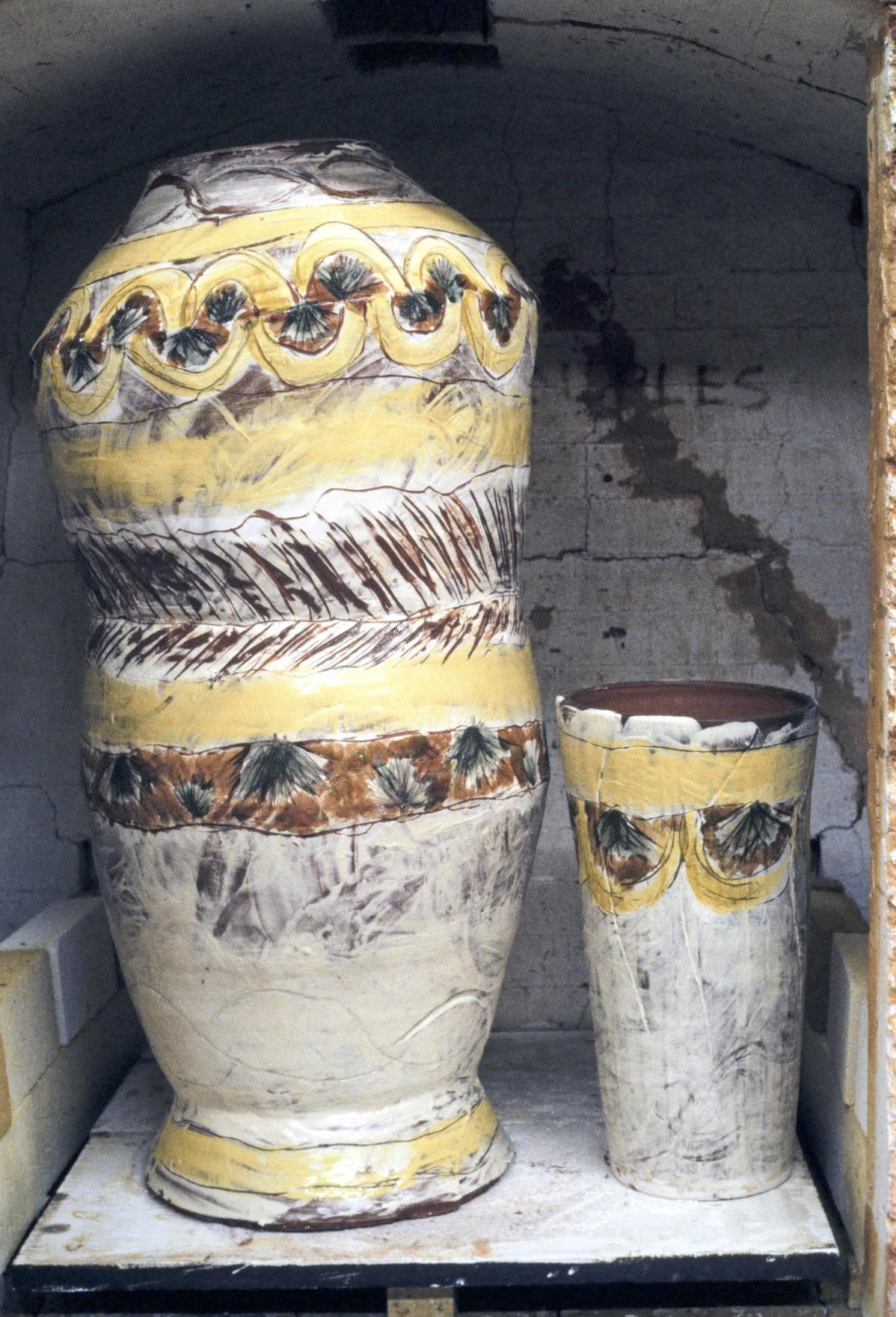 Two ceramics rest side by side in a kiln. The one on the left is a 4 foot tall sinuous figure-like vessel with brown and yellow patterning circling its white painted body. The one on the right is a foot tall in a loose cone shape with correlating patterning around the top lip. 