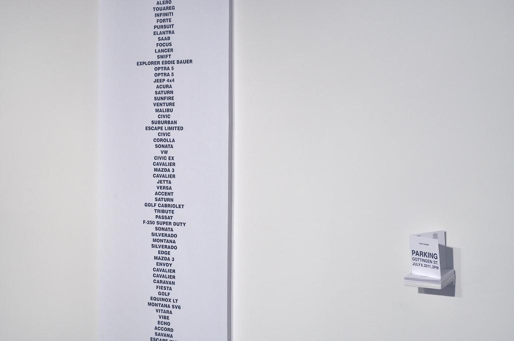 A tiny shelf holds paper folders which read: PARKING GOTTINGEN ST. JULY 8, 2011, 2PM. To the right hanging on the white gallery wall is a long strip of white paper stretched from floor to ceiling listing car names in black sans serif capitals: ALERO, TOUAREG, INFINITI, FORTE, PURSUIT, ELANTRA, SAAB, FOCUS, LANCER, SWIFT, EXPLORER EDDIE BAUER, OPTRA 5, OPTRA 5, JEEP 4X4, ACURA, SATURN, SUNFIRE…which continue out of sight. 