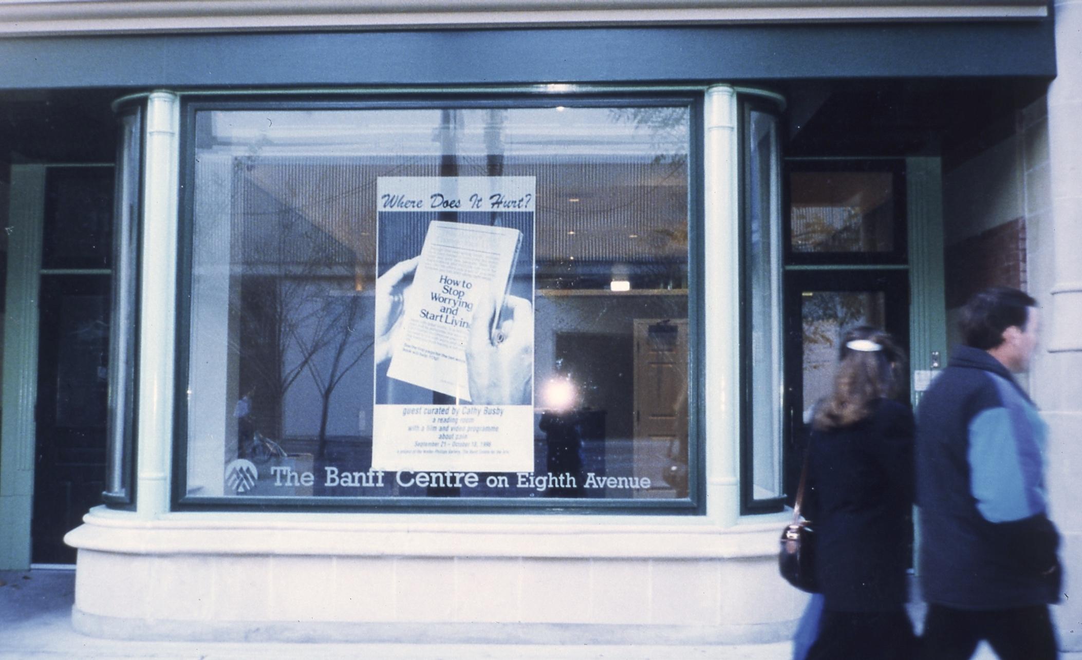 Two people hustle by the large bay window of the Banff Center off-site. A large poster hangs in the window: Where does it hurt? Is written in cursive script above a picture of two hands holding a book entitled: How to stop worrying and start living. The bottom half of the poster is blurry and obscured by the camera flash glaring off of the glass. 