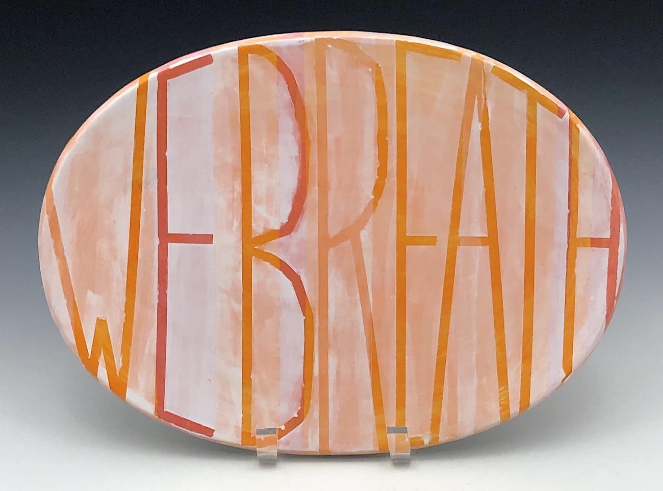 An ovular ceramic platter painted in pink, orange and white rests on a stand. WE BREATH is written in thin tall lettering which bends and stretches, pressing against the borders of the plate at every edge. 