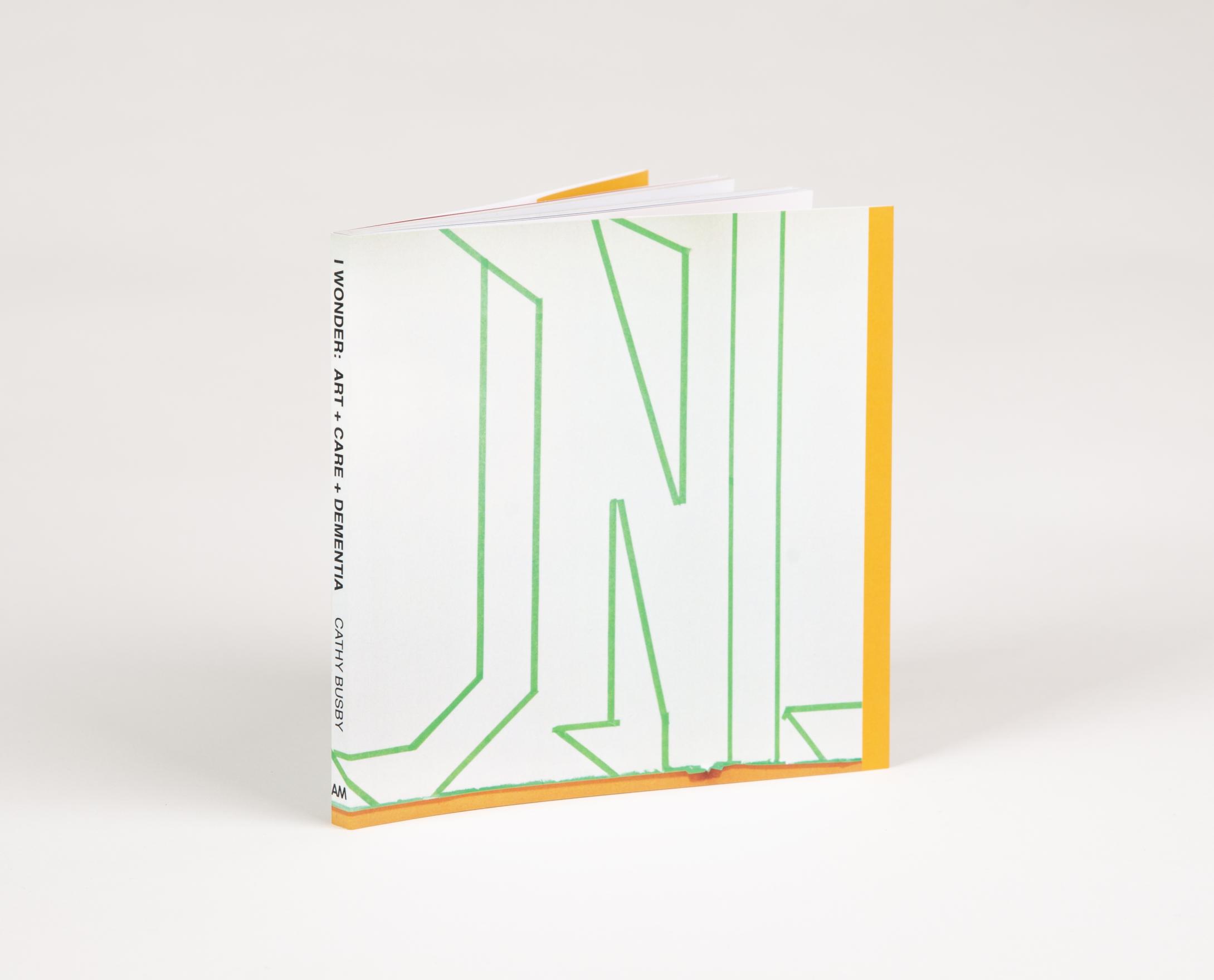 A small square book rests upright at an angle, the spine reads: I WONDER: ART + CARE + DEMENTIA CATHY BUSBY AM. The cover of the book is white with abstract letter-looking outlines in green painters tape. 