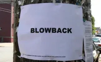 On a sunny street, a telephone poll speckled with staples and residue of past posters holds a fresh white piece of printer paper with the word BLOWBACK typed out in bold black sans serif font. 