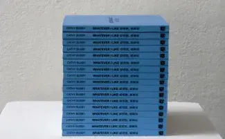 A stack of small blue publications lie on a white plinth. Each one’s spine reads: CATHY BUSBY, and WHATEVER I LIKE in both English and Mandarin. 