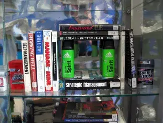 Self-help books and various brands of deodorant are assembled in a silver lined vitrine. The book have titles like: SELF-MANAGEMENT, WORKING WELL, MANAGE THE MIND, STRATEGIC MANAGEMENT, and EMPOWERED. Four neon green cans of ADIDAS deodorant hold up a stack of books like a roof of a house. 