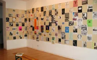A gallery wall is filled with a chess board layout of posters on a beige background. The posters are all different shapes, sizes and colour combinations, some look hand drawn others are typed. None are readable from this distance.