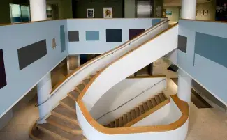  A curving wooden staircase leads between floors of a gallery with a bright modern interior. Light blue walls feature rectangular color-block silhouette wall paintings in various blue and gold tones. The upper level of the gallery displays framed art.