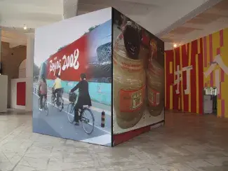 A cube made of four large panels of stretched vinyl rests in the centre of a high ceiling gallery space. The panel on the left holds a picture of cyclists riding past a 2008 Beijing Olympics sign, the panel on the right is an enlarged image of sauces on a Chinese grocery store shelf. 