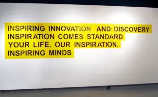 A white gallery wall holds a vibrant wall text painting. Black stenciled lettering on a yellow background reads: INSPIRING INNOVATION AND DISCOVERY INSPIRATION COMES STANDARD YOUR LIFE. OUR INSPIRATION. INSPIRING MINDS