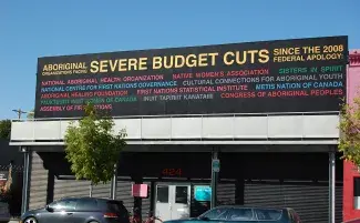 A large black billboard looms atop a roadside gallery space. The multi-coloured text which fills the sign reads: ABORIGINAL ORGANIZATIONS FACING SEVERE BUDGET CUTS SINCE THE 2008 FEDERAL APOLOGY: NATIONAL ABORIGINAL HEALTH ORGANISATION, NATIVE WOMEN’S ASSOCIATION, SISTERS IN SPIRIT, NATIONAL CENTRE FOR FIRST NATIONS GOVERNANCE, CULTURAL CONNECTIONS FOR ABORIGINAL YOUTH, ABORIGINAL HEALING FOUNDATION, FIRST NATIONS STATISTICAL INSTITUTE, METIS NATION OF CANADA, PAUKTUUTIT INUIT WOMEN OF CANADA...