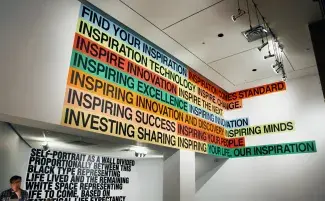A mezzanine wall is painted with a colourful wall-text painting. Each phrase is painted in black capital letters on either eggshell blue, bright red, cucumber green or light yellow.The phrases read: FINE YOUR INSPIRATION. INSPIRATION COMES STANDARD. INSPIRATION TECHNOLOGY. INSPIRE CHANGE. INSPIRING EXCELLENCE. INSPIRING INNOVATION. INSPIRING INNOVATION AND DISCOVERY. INSPIRING MINDS. INSPIRING SUCCESS. INSPIRING YOUR PEOPLE. INVESTING SHARING INSPIRING. YOUR LIFE, OUR INSPIRATION. 