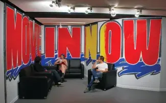 Three people sit on black couches in a break room. A large wall-text painting which reads: MOVE IN NOW in bright red and yellow lettering overshadows their bodies. The painting is done in a graffiti like style with blue marker scribbles scribed below the phrase. 