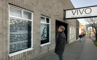 A man stands on the street looking at a window art installation at VIVO Media Arts Centre. One window is filled by a vinyl cut out of an Indigenous bird artwork, the other is filled with text, the vinyl lettering which is only partially legible and reads: ELDER HARRIET NAHANEE…DID NOT APOLOGISE…FOR HER CONDUCT…INDIGENOUS RIGHTS…LAND USED…SEA-TO-SKY HIGHWAY FOR…OLYMPICS. HARRIET NAHANEE WAS…JANUARY 24, 2007 AND…HEALTH, IMPRISONED FOR 14…THE SURREY PRE-TRIAL…DIED SHORTLY AFTER HER RELEASE…