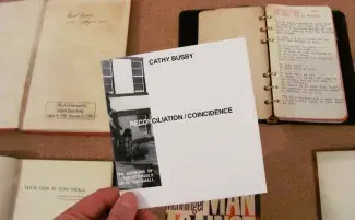 Fingers pinch the corner of a thin paper folder. The cover is white with a cropped black and white image stretching around from the back. CATHY BUSBY is written in black capital letters at the top and the title: RECONCILIATION/COINCIDENCE lies in the middle. Below the featured folder are various old looking books opened on display. 