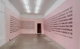  Three walls of a cement floored gallery are painted in two tones of light pink. Black capital lettering fills the walls but is mostly illegible, fragments read: WE SAY SORRY TO THE MOTHERS AND…FAMILIES…PARLIAMENT OF AUSTRALIA…IN THE SPIRIT WHICH IT IS…