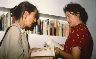 Cathy Busby, wearing a red floral dress with her brown hair pinned back, speaks with Moyra Davey, wearing a cream sweater and her hair in a bun. The two are holding open books standing in front of a single bookshelf built into the white gallery wall. 