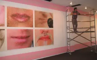 A white gallery wall is filled by a grid of 4 large photo prints of mouths. These closely cropped images are blown up large so the individual dots in the halftone pictures show. Each mouth is frozen either just about to speak or pursed shut in a word swallowing frown. The top and bottom of the wall is lined with a pink frieze with pale skin toned text which can be partially read: apologise to everyone, including my family. The rest of the text fades behind a man standing on floor to ceiling scaffolding.