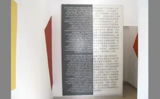  Fabric panels hang in a gallery space with yellow and red shapes on the walls. White text on a grey panel is written in English and grey text on a white panel is written in Mandarin, both texts are barely legible with font size and image quality. 