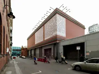Glancing up from an urban laneway, a large brick building is partially wrapped in two billboard sized vinyl sheets coloured pinky-white like pale blushing skin tone. This vinyl is covered in bold black text which is unreadable at this distance but the scale of the signs draws the attention of the few people passing by. 