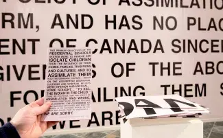A hand holds up a pink pamphlet with bold black text in front of a matching wall vinyl where the text is blown up and stretched from floor to ceiling. There is also a white plinth with a stack of pink vinyl fragments, remnants of another panel cut up for viewers to take. The leaflet reads in-part: TWO PRIMARY OBJECTIVES OF THE RESIDENTIAL SCHOOLS SYSTEM WERE TO REMOVE AND ISOLATE CHILDREN FROM THE INFLUENCE OF THEIR HOMES, FAMILIES, TRADITIONS AND CULTURES, AND TO ASSIMILATE THEM INTO DOMINANT CULTURE…