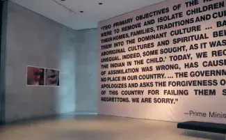 A large pink banner with bold black text stretching from floor to ceiling looms over the high ceiling concrete atrium. There are two closely cropped photos of mouths mid-way through speaking that hang on the wall adjacent. The banner is partially readable: “TWO PRIMARY OBJECTIVES OF…WERE TO REMOVE AND ISOLATE CHILDREN…THEIR HOMES, FAMILIES, TRADITIONS AND CUL…THEM INTO THE DOMINANT CULTURE…ABORIGINAL CULTURES AND SPIRITUAL..UNEQUAL. INDEED, SOME SOUGHT…THE INDIAN IN THE CHILD.’ TODAY, WE…OF ASSIMILATION...
