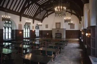 The grand refectory hall in the Union Theological Seminary is empty except for rows of dark wood tables and chairs. The vaulted ceilings are illuminated by candelabra chandeliers and large brown squares fill voids where paintings once hung.
