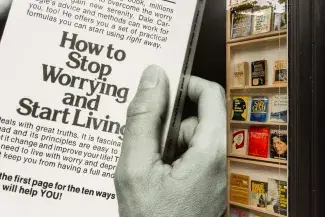 A black and white picture of a hand holding a book entitled: How to Stop Worrying and Start Living, fills most of the gallery front window. Sticking out from behind the large photo poster is is a bookshelf lined with front facing self-help books, some of their titles read: Sad Glad, Supermom, Sexual Suicide, The Pain of Being Human, Perscriptions for Anxiety, The Booze Battle.