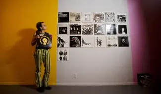 Cathy Busby stands against a bright yellow wall wearing green striped pants holding a Power in the Darkness vinyl with a drawing of a fist in the same yellow. She looks up to her left at a wall full of vinyl records with black and white album covers installed in rows on the wall. 
