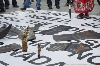Sacred objects made of copper, rock and feather lie on top of a pink vinyl poster covered with bold black sans serif text. The text is unreadable, obscured by both the objects and a pair of feet in black flip flops. Only this person’s legs and feet are in the frame which are covered in a decorated red robe. The feet of onlookers line the concrete perimeter of this display, each standing still or shifting their weight, waiting to see what the performer or the objects do next.