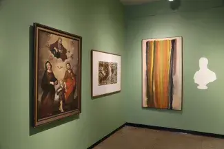 The corner of a gallery room, painted in avocado green, is lined with three artworks and the white shadow of a bust painted onto the wall in white. The paintings are to the right of the bust: a tall water colour of purple, yellow, green and orange stripes, then a black and white piece which looks like a lithograph, and a renaissance style painting of the holy family on the far left.