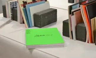 A neon green book rests on a white table. Attitudes 2017 is scribbled in black cursive across the front. There are black bricks behind this book which are acting as bookends for an array of thin publications in white and blue and black with no clear titles, just unreadable snippets of red or black text. 
