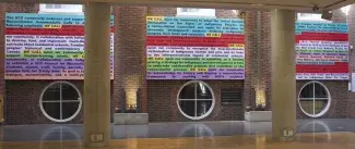The words WE CALL, written in black text and highlighted in yellow, punctuate three fabric wall-text panels where blocks of text are highlighted in blue, red, purple, green and pink. These panels are hanging in an atrium of a brick walled college campus above three circular windows. The text is slightly obscured by two concrete pillars but partially reads: The UCC community endorses and supports…Reconciliation Commission’s Call to…following emphasis: WE CALL upon our…