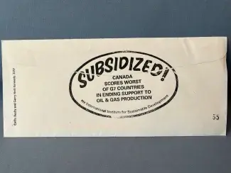 A white business envelope lies face down. A black oval stamp encircles the sealed closure, contoured to the shape of the stamp the words read: Subsidized! Canada scores worst of G7 countries in ending support to oil & gas production. See International Institute for Sustainable Development. Another stamp runs vertically up the edge of the envelope which says: Cathy Busby and Garry Neill Kennedy, 2019.
