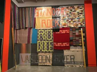 A patchwork of plastic, tape and multi-colour knitted and quilted blankets fill the high wall of a gallery space creating a sense of both urgency and utility with their activist signage aesthetics. Near the top of the wall, orange tape on clear plastic spells out: STANDING ROCK, below, taped directly on the blankets, in lime green: RIO DOCE, and close to the floor, in grey: WABIGOON RIVER. Leaning against the wall are 2 by 4’s wrapped in cloth, ready to be wielded or moved as the situation requires. 