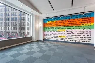 The words WE CALL, written in black text and highlighted in yellow, punctuate a wall-text painting where blocks of text are highlighted in blue, orange, green, light pink and cream. Adjacent to the painted wall is a set of floor to ceiling windows overlooking Downtown Vancouver, the rest of the room is empty. The text reads in part: United Nations Declaration on the Rights of Indigenous peoples, Treaties and Aboriginal rights, and Indigenous law. WE CALL upon governments in collaboration with Survivors…